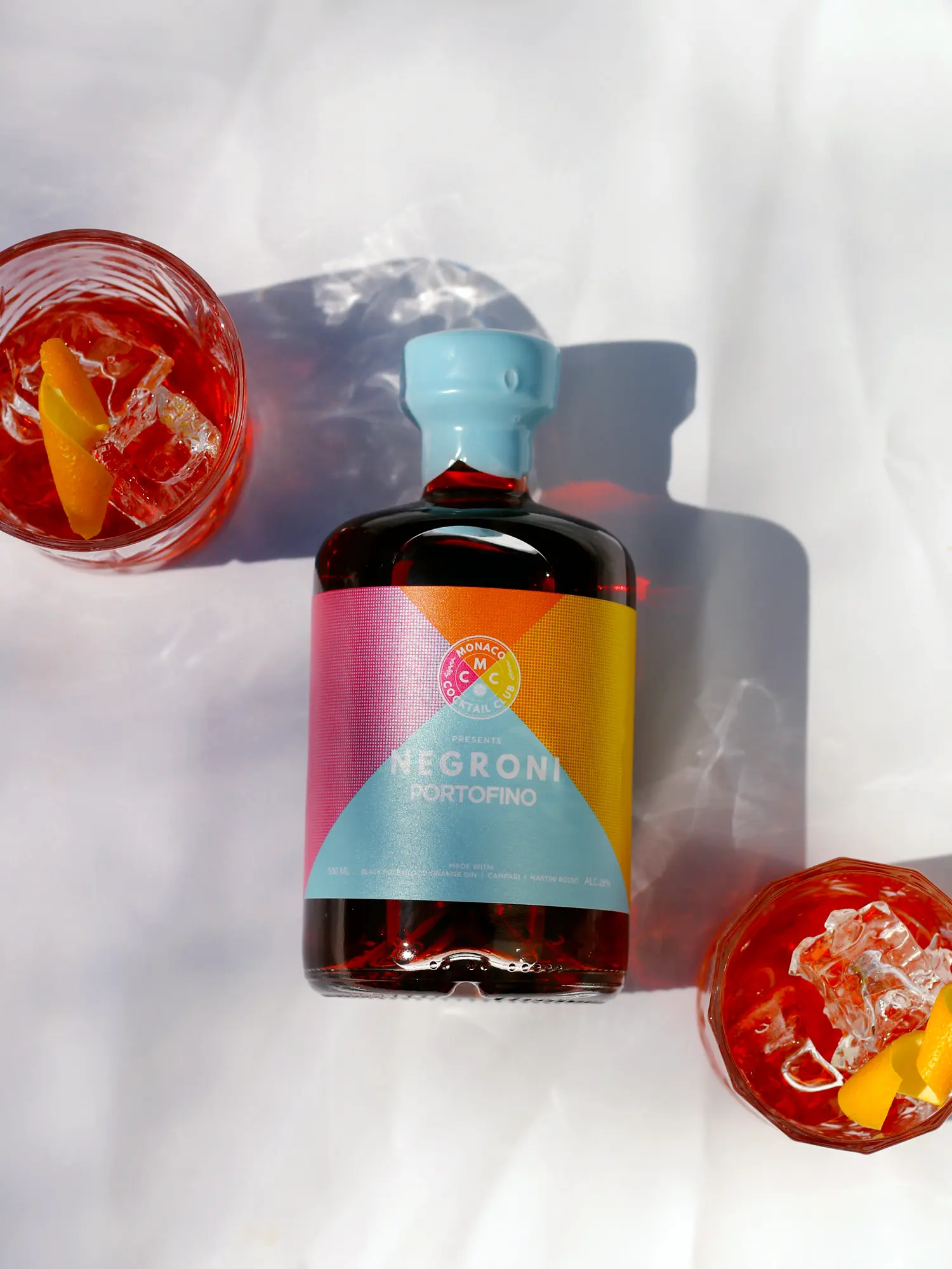 Negroni Portofino Bottle and Serves - Top Down View A modern flat lay of the Monaco Cocktail Club's Negroni Portofino bottle, artfully arranged between two prepared cocktails in crystal glasses. The scene, shot on a bright white surface with harsh sunlight creating beautiful shadows, highlights the vibrant colors of the product.